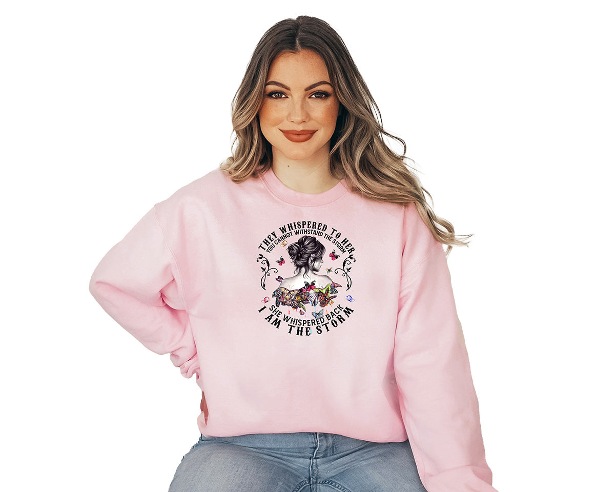 Woman wearing a baby pink sweatshirt with a big screen printed. The silhouette of a young lady's back with small colorful butterflies around her and a text around the design they whispered to her you cannot withstand the storm.