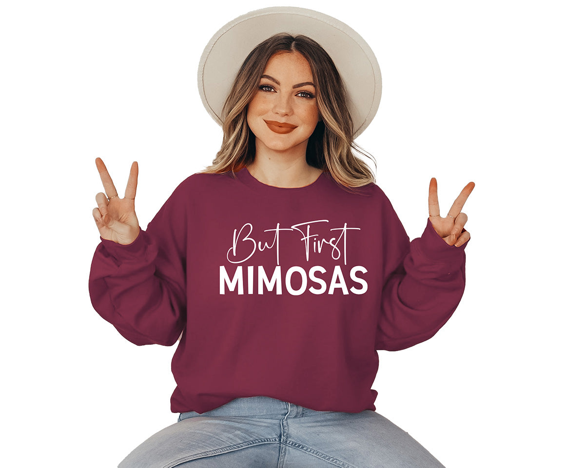 Woman wearing a garnet sweatshirt. On the front of it there is a big white screen printed in Calligraphy says But First and in Caps Mimosas.