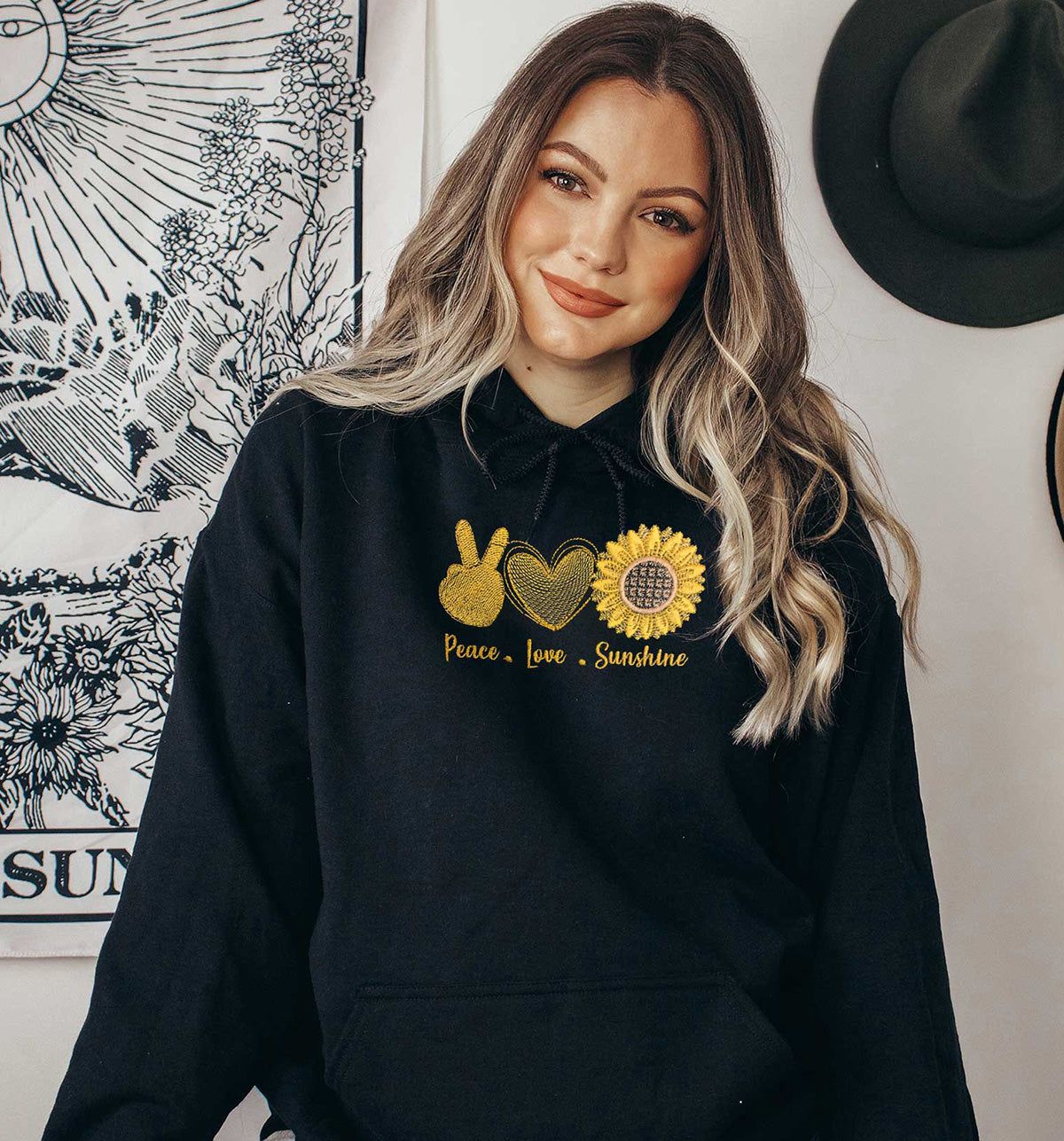 Young lady wearing a black hoodie. The yellow embroidered design has three images, the sign of peace, a heart and a sunflower. Below the images a text of Peace Love Sunshine