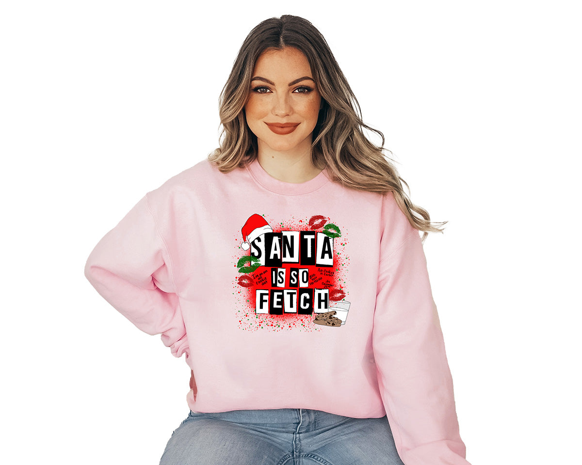 Woman wearing a light pink crewneck. This one has a large red screen printed of Christmas. A cap hanging in the corner of the white and black text Santa is so fetch. Green and red kisses images around the text and a glass of milk and chocolate cookies at the end of the text  