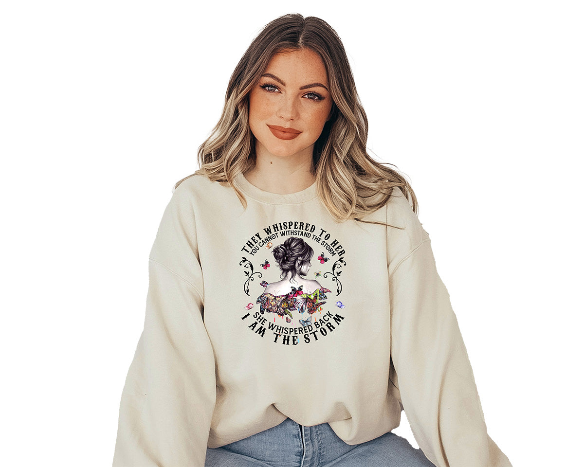 Woman wearing a sand sweatshirt with a big screen printed. The silhouette of a young lady's back with small colorful butterflies around her and a text around the design they whispered to her you cannot withstand the storm