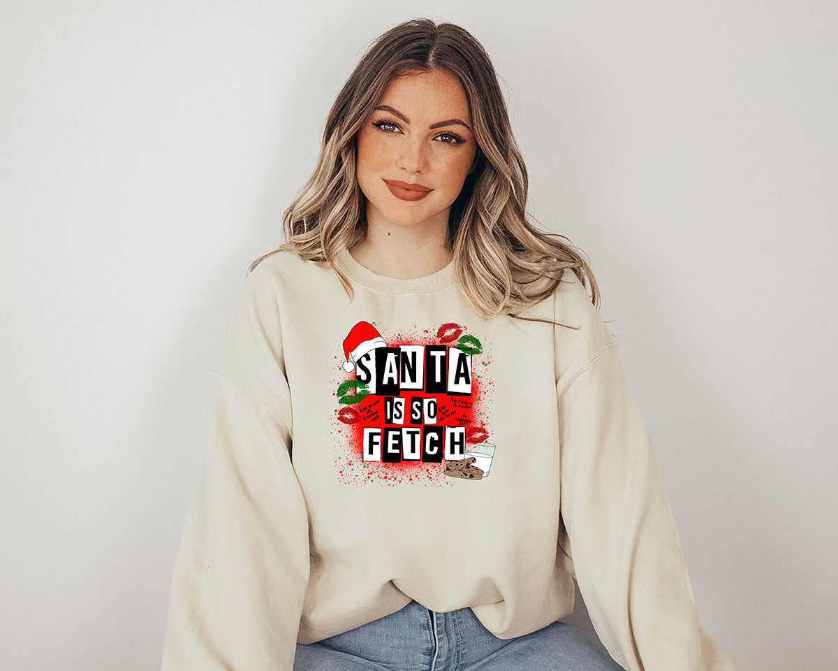 Woman wearing a sand crewneck. This one has a large red screen printed of Christmas. A cap hanging in the corner of the white and black text Santa is so fetch. Green and red kisses images around the text and a glass of milk and chocolate cookies at the end of the text.