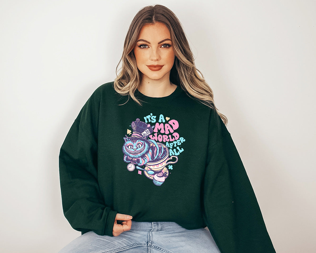 Young lady wearing a forest green crewneck sweatshirt with a screen printed. A big colorful smiley cat with a text it is a mad world after all.