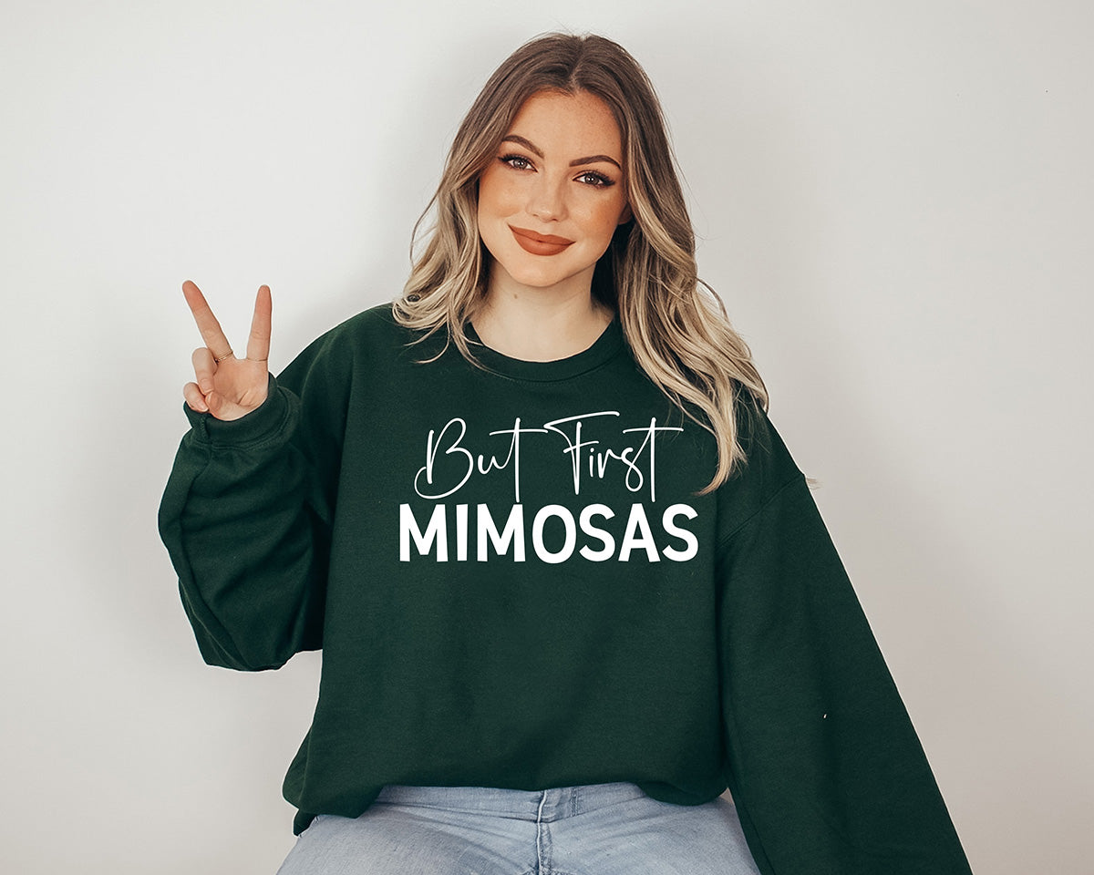 Woman wearing a forest green sweatshirt. On the front of it there is a big white screen printed in Calligraphy says But First and in Caps Mimosas.