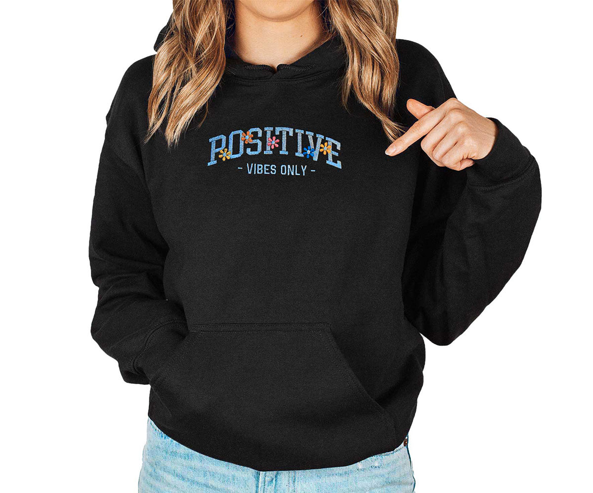 Young lady wearing a black hoodie. Positive Vibes only is embroidered in turquoise thread color and colorful little flowers around the text.