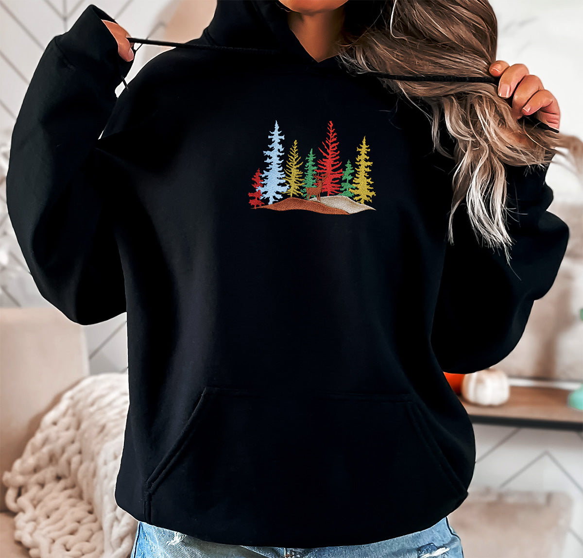 Young lady wearing an embroidered black hoodie. She has a design of seven pines, they are in different colors, two red. two green, two mustard and one light turquoise. In front of the seven pines there is a brown deer standing.