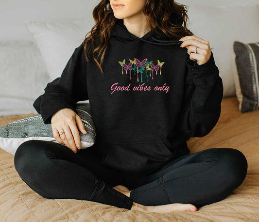 Young lady wearing a black embroidered hoodie. Aligned butterflies, pink, yellow and green colors. Below there is a embroidered design text in calligraphy Good Vibes only in color pink.