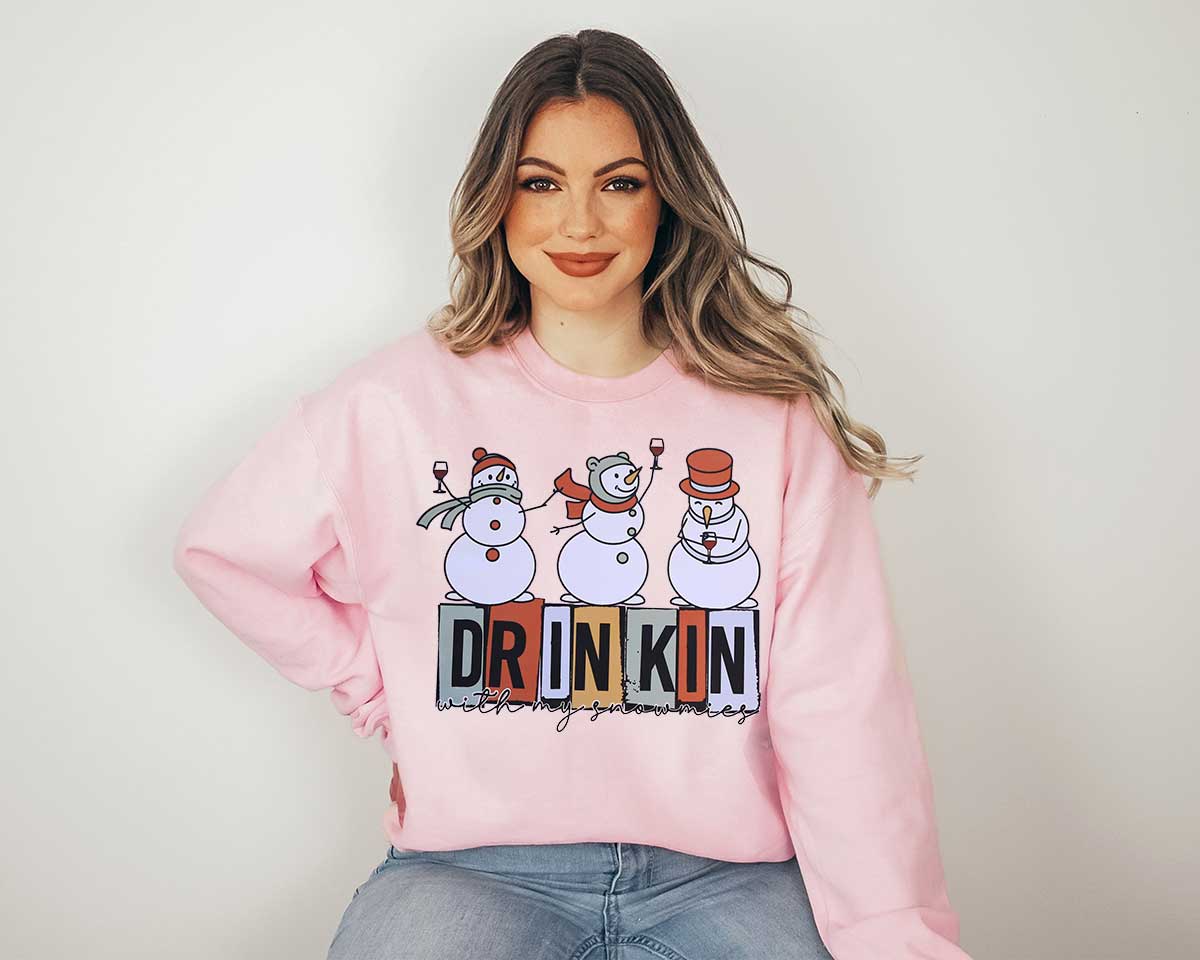 A woman wearing a light pink sweatshirt with a big screen printed of three snowmen drinking wine with the logo drinking with my snowmies