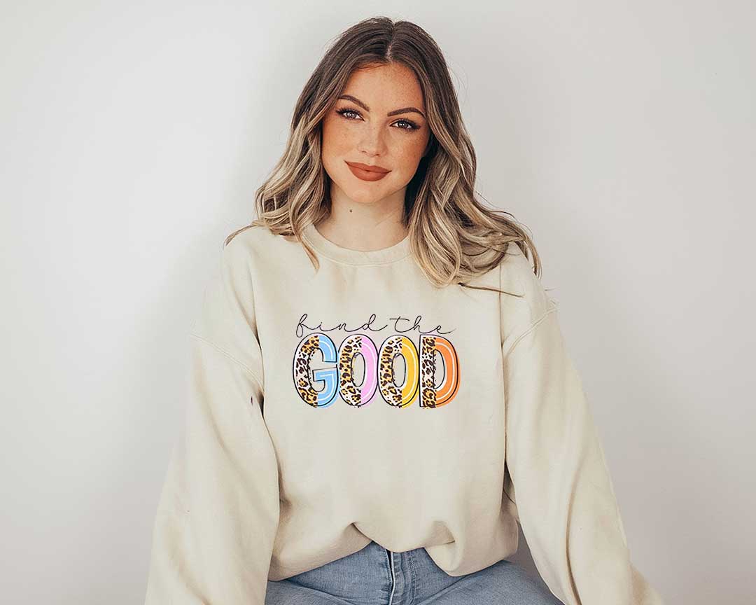 Woman wearing a sand sweatshirt with a big screen printed on the front. In black calligraphy letters find and in large colored letters  Good