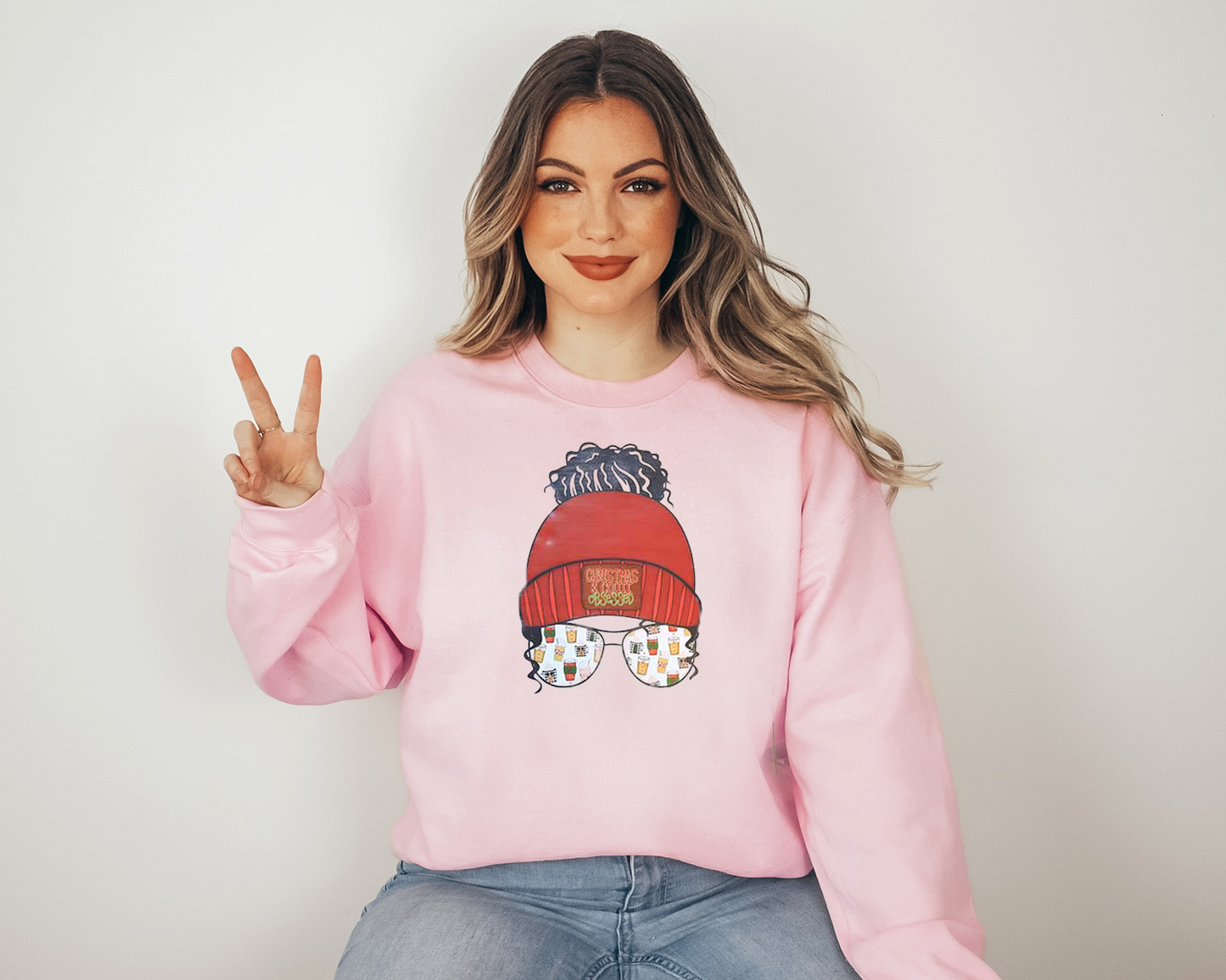 A woman wearing a light pink sweater with a big screen printed on the front. A red beanie with a logo on the cuff Christmas coffee obsessed. A black messy bun and images of cups of coffee in the sunglass. 