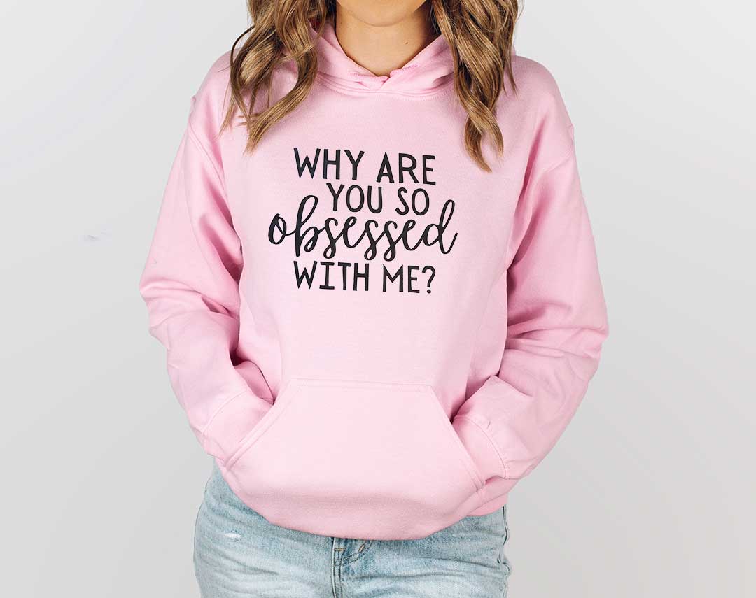 Young lady wearing a pink crewneck sweatshirt. Screen printed transfer in big black letters on the front. Why are you so obsessed with me?