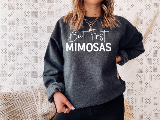 Woman wearing a dark heather sweatshirt. On the front of it there is a big white screen printed in Calligraphy says But First and in Caps Mimosas.
