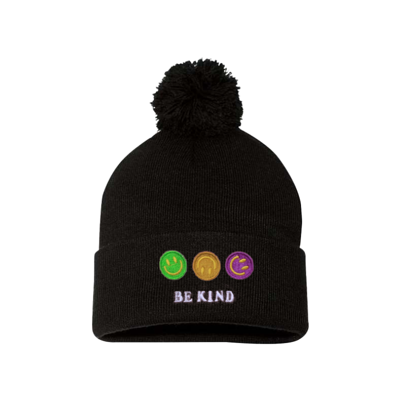 Black cuffed beanie with a pom pom. It has an embroidery on the cuff of three happy faces, green, gold and purple and a text in caps Be kind