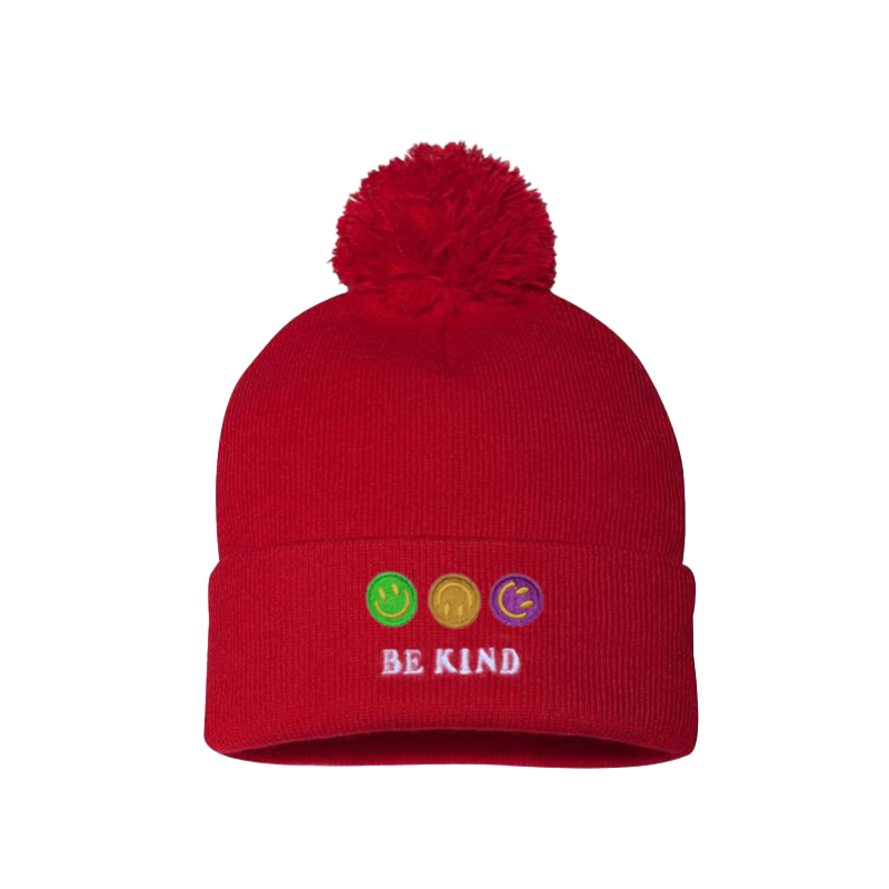 Red cuffed beanie with a pom pom. It has an embroidery on the cuff of three happy faces, green, gold and purple and a text in caps Be kind 