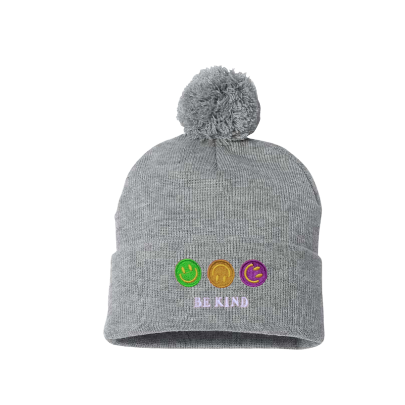 Light Gray cuffed beanie with a pom pom. It has an embroidery on the cuff of three happy faces, green, gold and purple and a text in caps Be kind