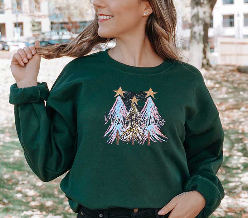 Young lady wearing a forest green sweatshirt with a large screen printed. Three Christmas trees with colors of blue and pink and a text of merry Christmas