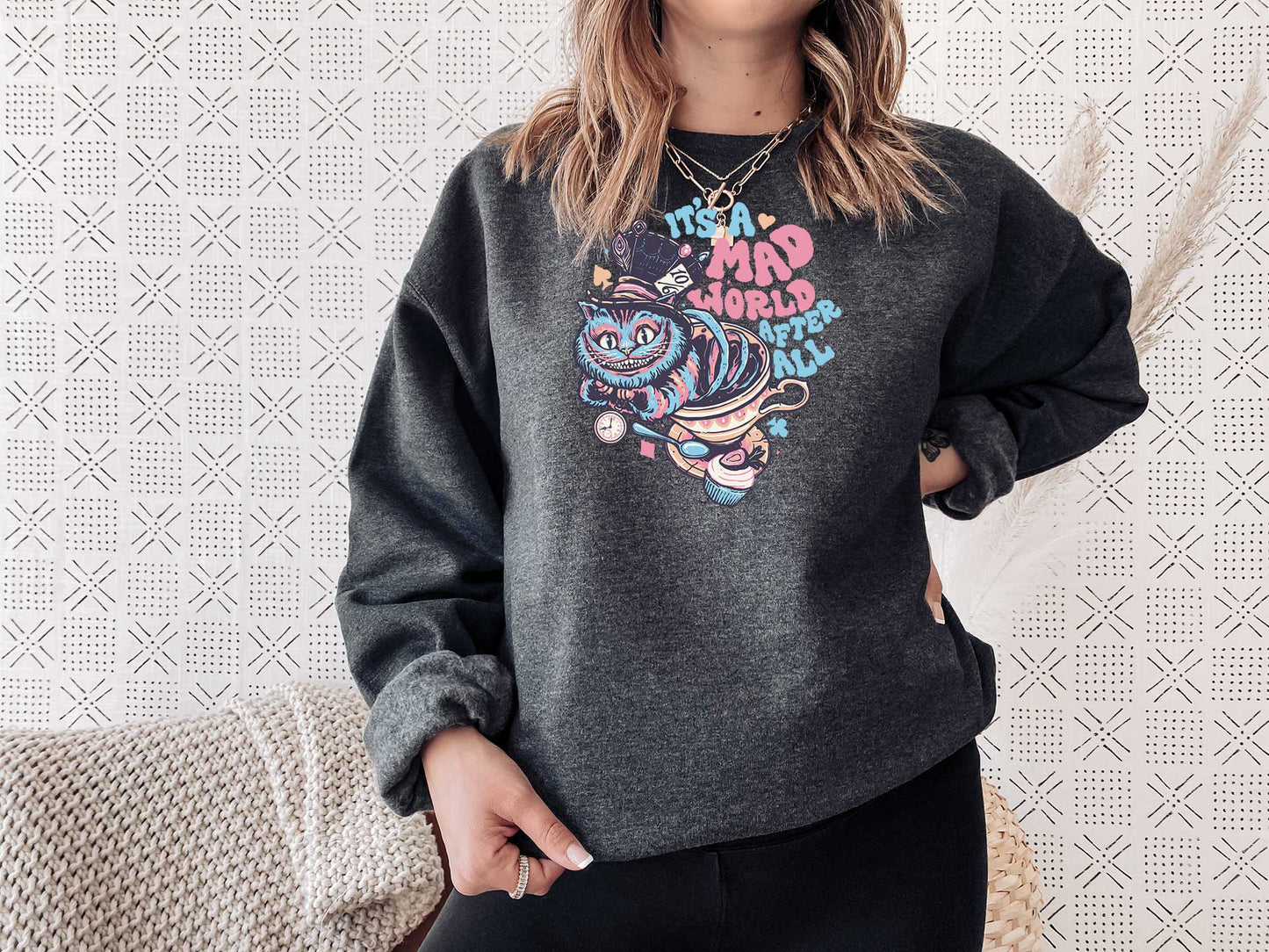 Young lady wearing a grey heather crewneck sweatshirt with a screen printed. A big colorful smiley cat with a text it is a mad world after all