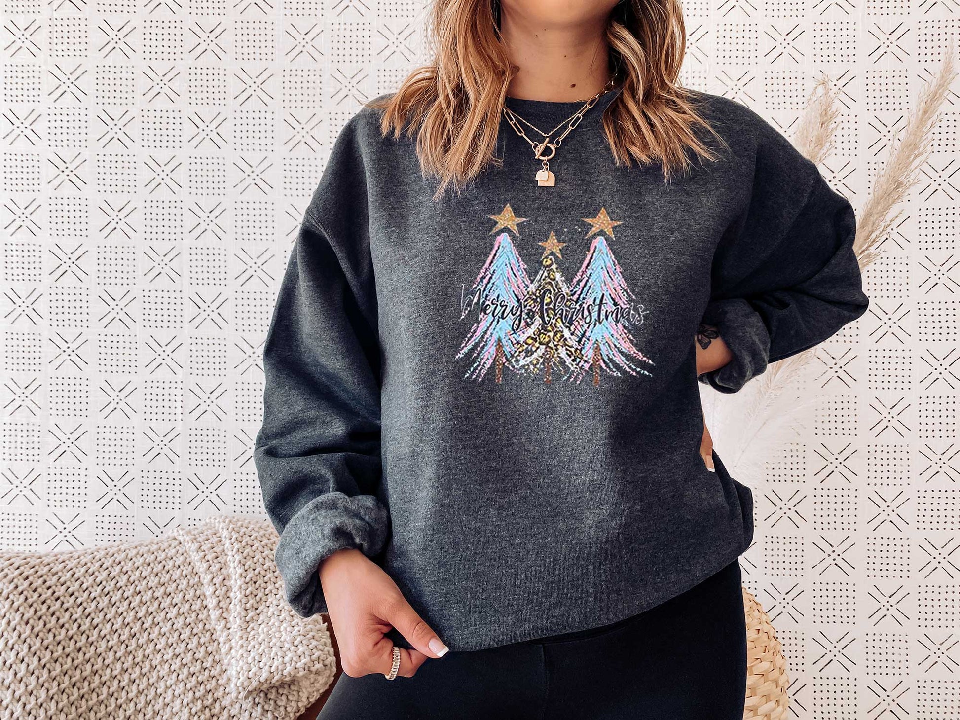 Young lady wearing a dark heather sweatshirt with a large screen printed. Three Christmas trees with colors of blue and pink and a text of merry Christmas